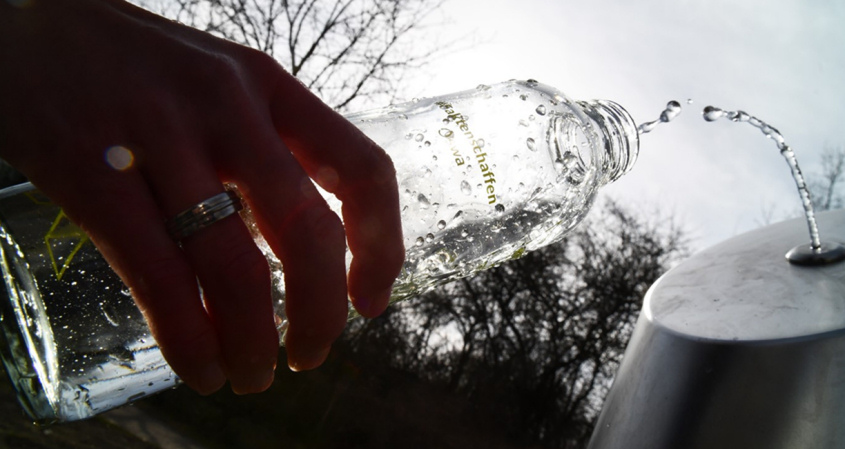 Hand hält eine Glasflasche unter einen Trinkwasserstrahl an einem öffentlichen Wasserspender im Außenbereich.
