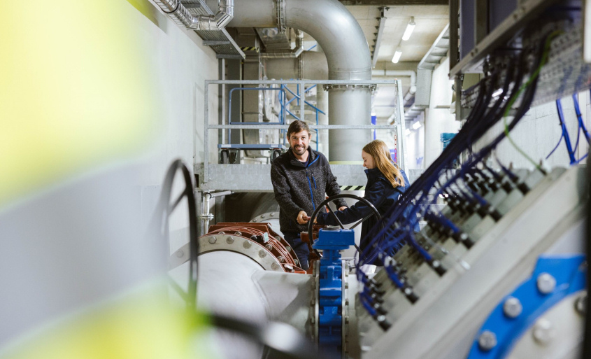 Technischer Anlagenraum mit großen Rohrleitungen und Mess- sowie Steuertechnik, in dem zwei Personen an einer Armatur stehen.